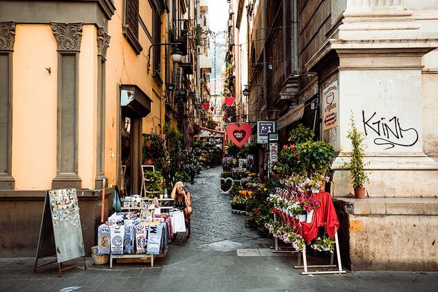 A street corner in Naples. A saleswoman sits outside her small general store, wiping her forehead with a handkerchief in the heat. Sales of flowers and souvenirs continue on into the alley. At the end of the alley, a restaurant can be seen.