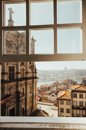 An open window looks out over a city by a river. In the foreground, just to the left outside the window, is the beautiful stone facade of a baroque church. Further away you see houses and rooftops, and on the other side of the river the city continues. The sky is blue, and it is sunny outside. What we see are parts of the city of Porto in Portugal.
