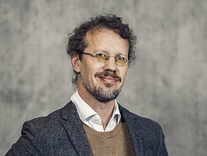 Johannes Persson, Dean of the HT Faculties, is photographed against a grey background. He has dark curly hair, a short beard and a moustache. His hair is in a ponytail. He is wearing glasses, and is dressed in a white button-down shirt and a brown sweater under a dark grey blazer.