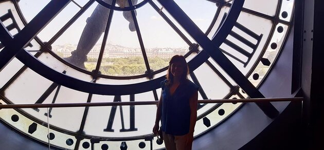 Paola is standing inside a building which houses a very large clock. Part of the clock work is visible behind her, and outside the window there is a view over Paris, with the white church Sacré-Coeur in the far background.