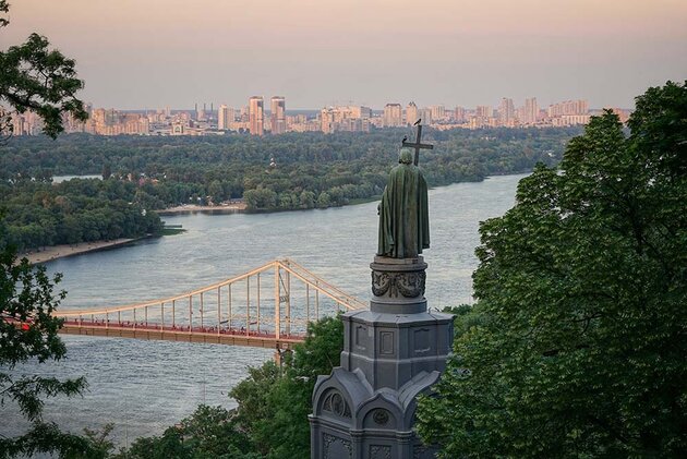 En vy över Ukrainas huvudstad Kiev. I förgrunden en bronsstaty som blickar ut över floden nedanför. Över floden löper en hängbro, och på andra sidan floden syns stadens parker och hus. Det är grönska och träd i förgrunden. 