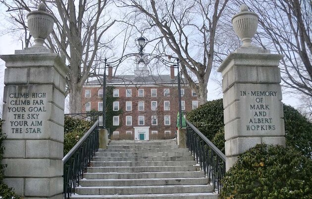 A stone staircase flanked by square columns with inscriptions leads up to a grand four-storey red brick building. A white bell tower can be seen on the roof of the building. On the roof of the building is a white bell tower. Several leafless trees are visible in the image; it appears to be winter in Massachusetts (we are looking at William's College).