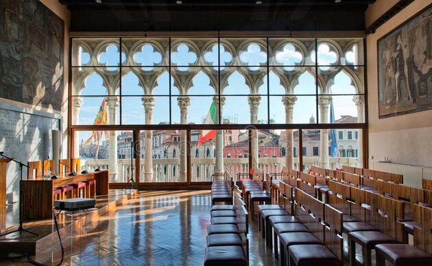 A view from inside a lecture hall in the Ca' Foscari University of Venice. Facing the canal is a large glass pane through which you can see the columns marking the window arches, three flying flags and also the rooftops on the other side. Inside the hall are rows of wooden chairs with brown leather seats and a podium with four chairs. Large paintings hang on the walls. The beautiful parquet floor is varnished and shines in the light from the window. 
