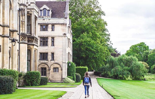 A young woman wearing a Fjällräven backpack walks on a cobbled walkway with her back to the camera. To her left are beautiful stone university buildings, to her right a well-manicured British park landscape. The picture was taken outside of King's College in Cambridge.