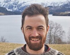 Greer is standing outside in front of a lake and a mountain, dressed in an Icelandic sweater. He has dark hair and beard and is smiling.