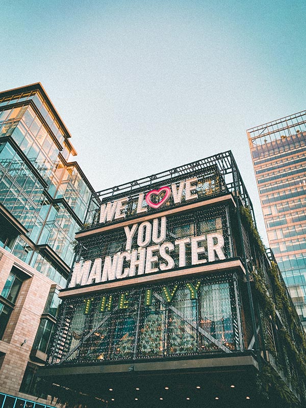 Three building facades are visible in the image. The one in the centre of the image has a large sign in the form of a three-line text, which can be illuminated in the evening. The text reads: “We love you Manchester”. The O in “love” is shaped like a red heart. Underneath, in other letters, it says “the Ivy”. The photo was taken outside The Ivy restaurant in Manchester.
