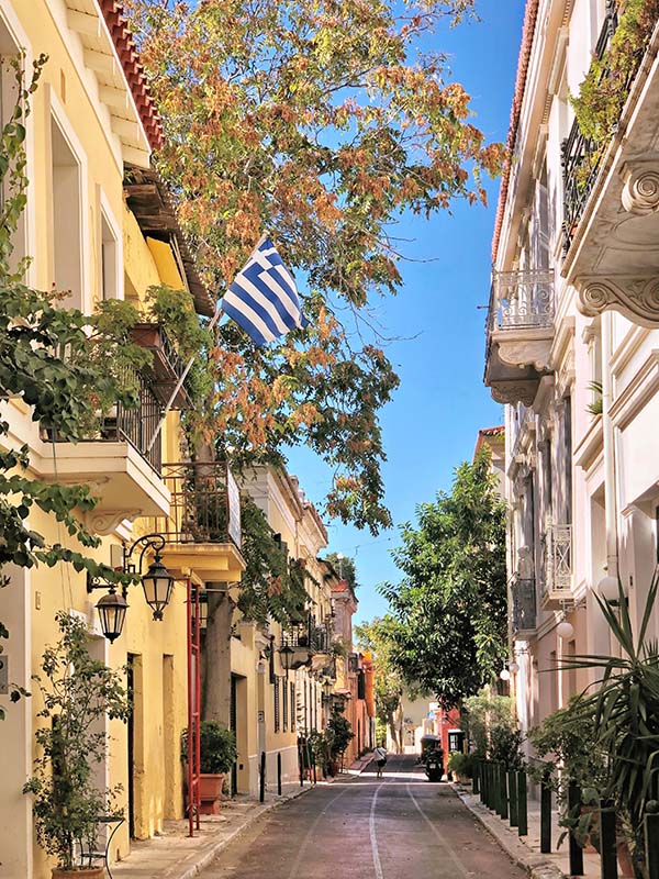 Bilden visar en lugn gata i Athen kantad av vackra hus i ljusa färger, med balkonger med snidade metallräcken. Långt fram på gatan vandrar en person med ryggen mot kameran. Träd kantar gatan och den grekisk flaggan vajar från en av balkongerna. 