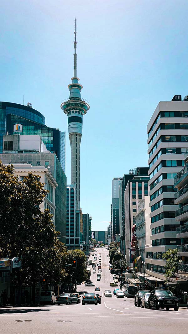 En gatubild från staden Auckland på Nya Zeeland. En flerfilig trafikerad gata leder in i bilden och kantas av höga flervåningshus. Längst bort tonar det höga tornet, Sky Tower, upp sig mot den blå himlen.
