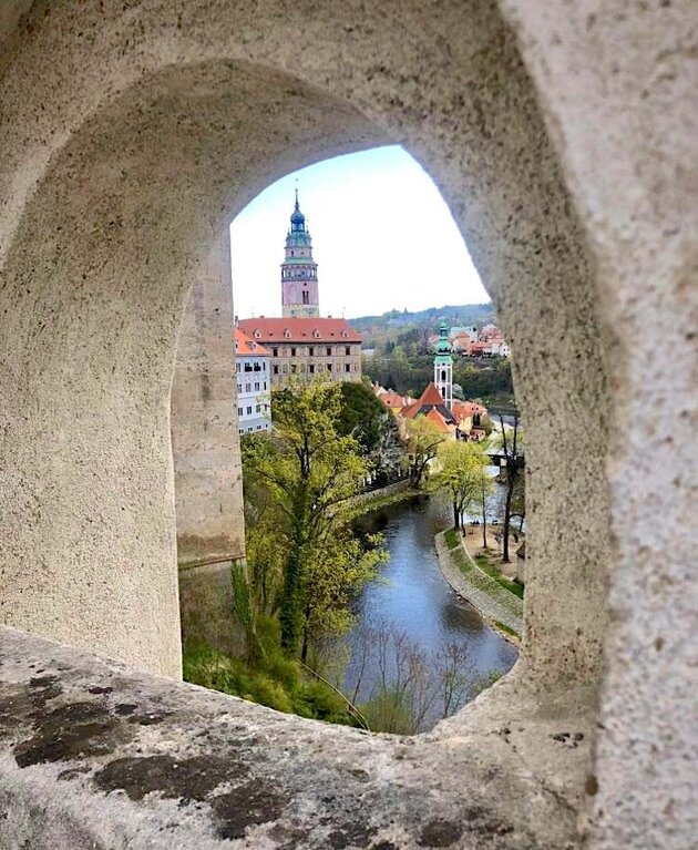 Through an arched hole in a grey stone wall, you can see a river, greenery and parts of a beautiful town with spires and towers. This is České Budějovice in the south of the Czech Republic.
