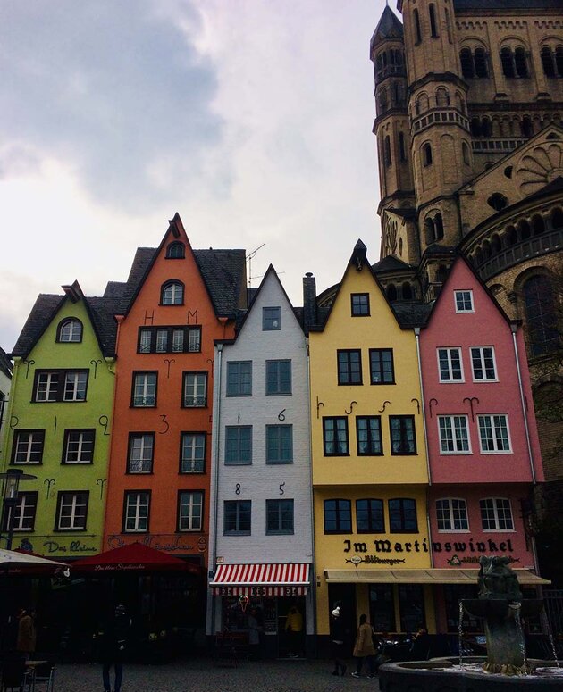 A square in Cologne, Germany, with a fountain in the foreground. On the other side of the square, there are pointed facades of beautiful old buildings of different colours. One of the buildings has the year 1685 written in metal numerals. At the top right of the image, a yellow-brick church can be seen in the background.