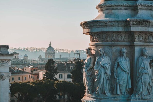Bilden är tagen i Rom. I förgrunden syns statyer som dekorerar en vit marmorbyggnad från 1800-talet, resten av bilden upptas av en vy ut över takåsarna i Rom. Längre bort syns en barockkyrkas kupol och längst bort i bilden syns kullar med pinjeträd.