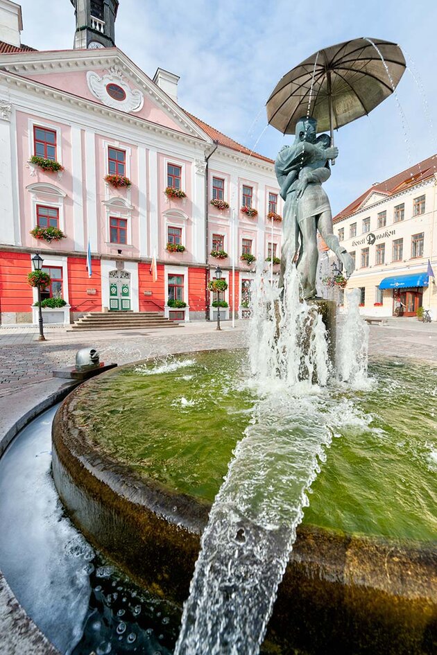 En bild från ett torg i den estländska staden Tartu. Vackert målade byggnader syns i bakgrunden och i förgrunden en vattensprutande fontän som visar en ung man och en kvinna i brons som kysser varandra under ett paraply.