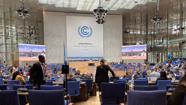 The image shows a large conference hall with blue-clad chairs in concentric rows surrounding a podium in the centre. Two large screens show a close-up of the people sitting on the podium. A large sign behind them reads “United Nations Climate Change Conference”. There are many people in the hall, mostly seated, but a man and a woman who have stood up to move around can be seen in the foreground.