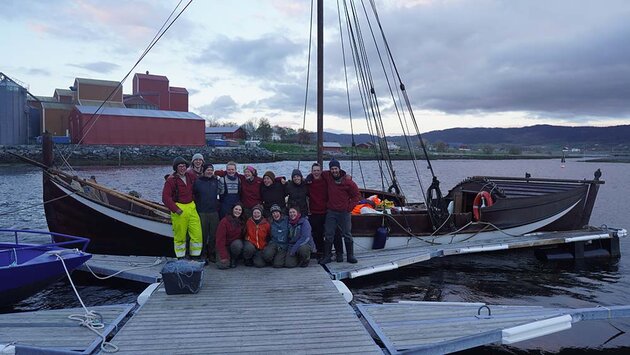 A wooden ship is moored at a pier in a harbour. In front of the ship, the crew, a group of 13 people, are smiling and looking happy after a successful voyage.