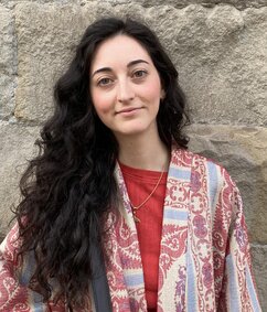 Caterina has long dark curly hair and glasses. She is wearing a jacket with colourful patterns.