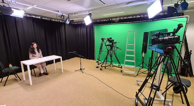 In a large studio with lights and recording equipment, Caterina sits at a white table, cross-legged. She has long dark hair and glasses, and is wearing a thick grey sweater and light-coloured trousers. There is a large green screen at the end of the room.