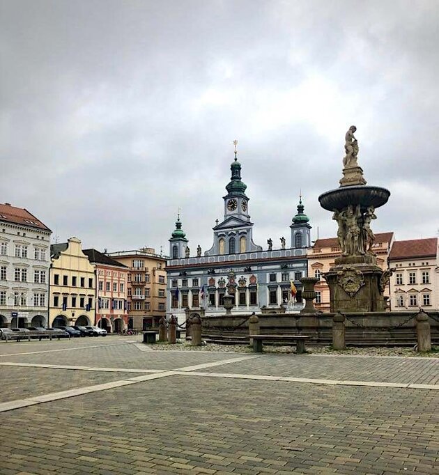 A cobblestone square houses a large ornamental fountain. Around the square are beautiful 18th and 19th century buildings in different colours. The square is located in the town of Český Krumlov in the south of the Czech Republic.