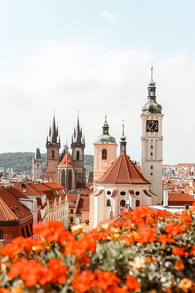 The image is taken over the rooftops of Prague, with orange flowers in a flower box in the foreground. The focus in the centre of the picture is on two churches with beautiful bell towers. A little further away are the rooftops of the city and in the far distance are green, wooded hills.