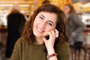 Luisa is looking at the camera. She has dark shoulder-length hair and brown eyes. Her head is tilted and resting on her left hand. She is wearing a green knit sweater. Luisa appears to be sitting in a bar or café. There are people standing and sitting at a counter in the background.