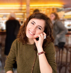 Luisa is looking at the camera. She has dark shoulder-length hair and brown eyes. Her head is tilted and resting on her left hand. She is wearing a green knit sweater. Luisa appears to be sitting in a bar or café. There are people standing and sitting at a counter in the background.