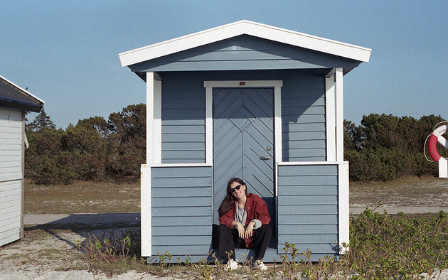 Luisa is sitting in front of a small blue beach house. She is wearing sun glasses. 