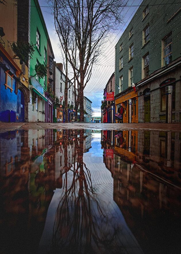 The image is divided into two parts. The upper part is a city scene, a street lined with colourful buildings, while the lower part is a reflection of the same view in a pool of water. Christmas decorations in the form of small Christmas trees with lights are visible on several of the facades. It is Christmas time in Galway, Ireland.