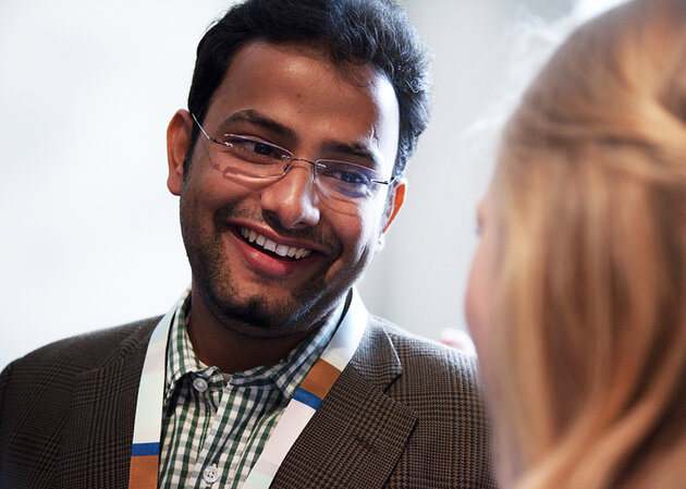 A meeting between two researchers. A man with short dark hair, beard and brown eyes smiles and looks at a woman with blond hair. The back of her head is visible in the right-hand side of the picture (rather blurry). The man wears glasses, a chequered shirt and a dark jacket. Around his neck there is a Lund University key band - he is in Lund at a conference.  