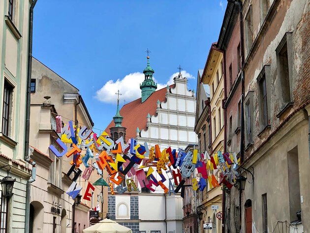 A city scene from Lublin, Poland. Between beautiful but worn building facades, rows of large colourful letters hang in a jumble. They appear to be made of stretched fabric. At the far end of the picture is a beautiful church-like building with a white facade with bronze details, a red-tiled roof and a bronze tower.