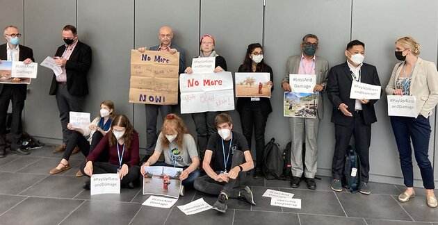 A group of twelve people are standing or sitting with placards in their hands They are demonstrating against something. The signs have text like “No more blah blah blah!” Several of them are wearing face masks. They are demonstrating during a climate conference. The four people sitting are young people dressed in jeans and T-shirts; the people standing are middle-aged people in suits or similar clothing.