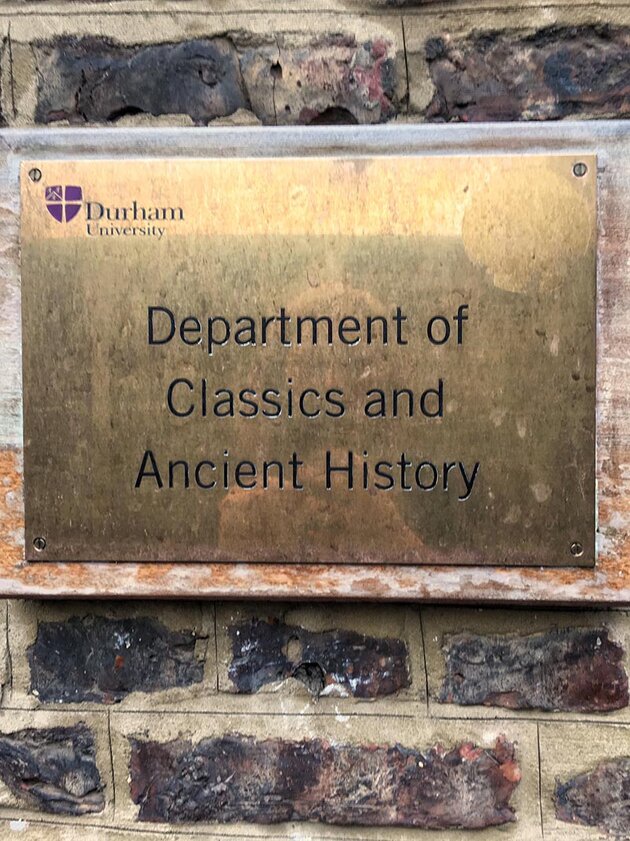 En guldfärgad metallskylt är uppsatt på en tegelvägg. På skylten står det ”Department of Classics and Ancient History”. Bilden är tagen vid Durham University i Skottland.