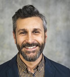 Nicolò has dark hair and beard, and a big smile. He is wearing a shirt and a grey jacket.