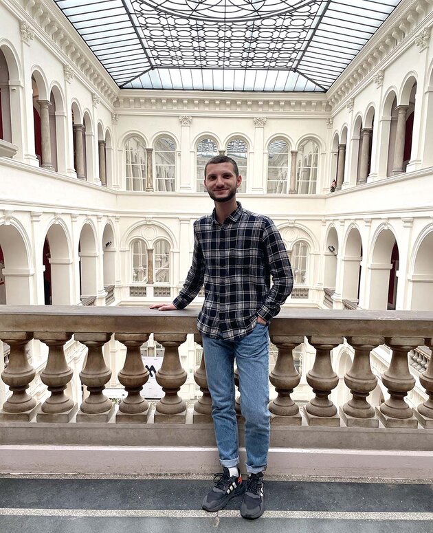 Uladzimir is standing in a white museum hall, dressed in jeans and a chequered shirt.
