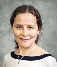 Ylva has her hair up, and is smiling. She is wearing a striped T-shirt under a blue and white sweater.