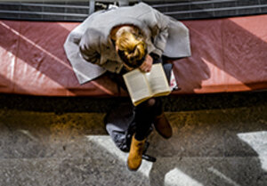En ung kvinna sitter på en bänk och läser i en bok. Bilden är tagen ur fågelperspektiv.