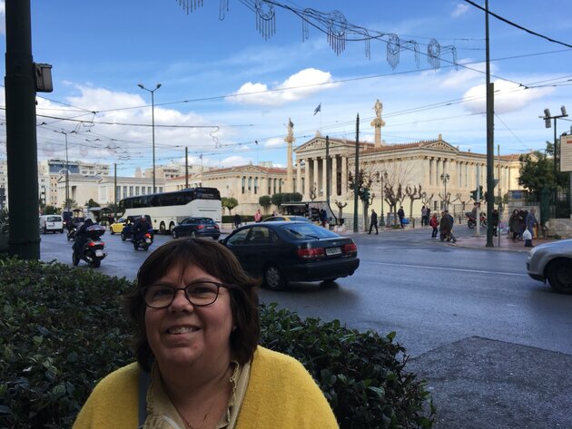 In the foreground, Malin is standing in front of a busy street. Malin is smiling and has dark, shoulder-length hair and glasses. She is wearing a yellow cardigan. Behind her, on the other side of the street, you can see a number of classical, beautiful buildings in Athens, including the main building of the university. 