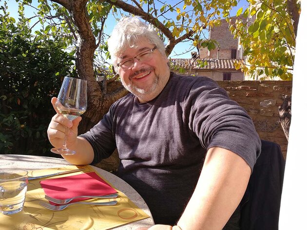 Stefan is sitting at a restaurant table smiling, holding up a glass with one hand. His hair is a bit messy from the wind. He has grey hair, a grey beard and moustache and glasses. He is wearing a dark blue shirt and has rolled up his sleeves. The sun is shining. Behind him, there is a stone wall, greenery and beautiful stone houses with tiled roofs. The picture was taken in San Gimignano, Italy.