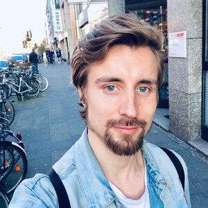 Juhan is standing on a city street. In the background, there are parked bicycles and people walking. He is wearing a light-coloured jeans jacket over a white T-shirt. The photo is a selfie. Juhan has brown wavy short hair, a dark beard and moustache and blue eyes. He is looking at the camera.