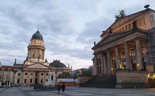 In an open space where only two people are moving, there are two majestic buildings: on the right, the Berlin Concert Hall with its column-lined entrance, and straight ahead, the beautiful Neue Kirche (New Church) with its round tower and bronze dome.