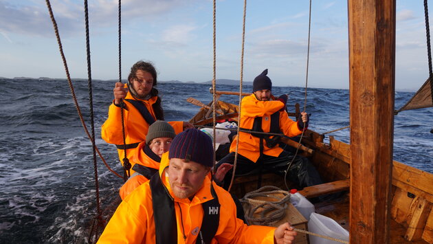 Four people are struggling with ropes hanging from the sails of an open wooden boat. They are all dressed in heavy orange rain jackets and life jackets. One of them, on the far right, is Greer. The sky is bright, but the sea looks dark and troubled. Land can be seen far away in the distance. 