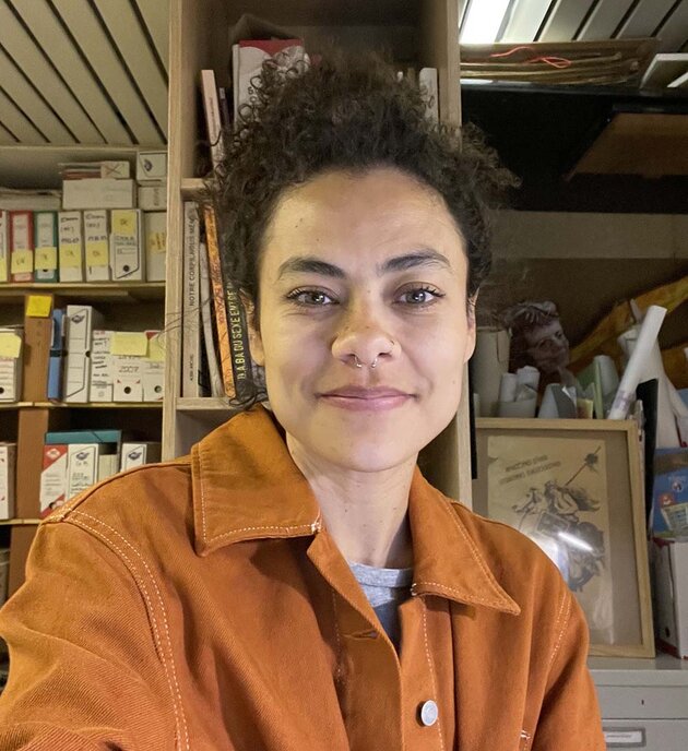 Camila is sitting inside the "Archives Lesbiennes" in Paris. She has her dark curly hair up, and is wearing a brown shirt.