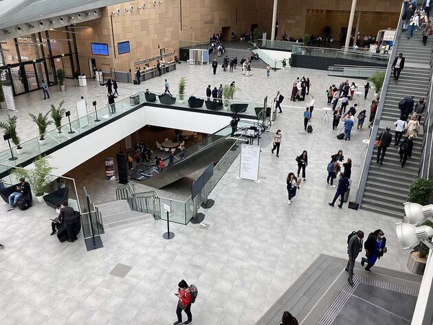 An atrium inside a conference building with stairs leading up and down and many people moving around. The picture is taken from an upper floor. Green plants and large signs are scattered here and there.