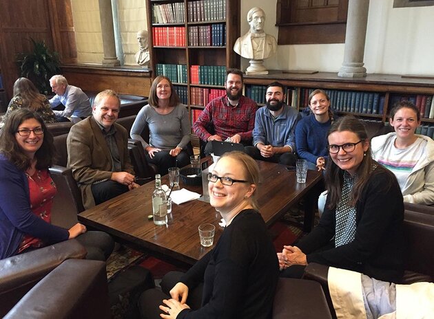 In a room that appears to be a library, with books and plaster busts of great thinkers, a group of nine people sit smiling and facing the camera. Located on the right side of the picture is Maria Småberg, with dark hair and dark glasses. Her hands are clasped in her lap, and she is wearing a patterned blouse and black cardigan. Beside her is her colleague Lina Sturfelt, her light-brown hair up wearing a black sweater. Both ladies are smiling happily.