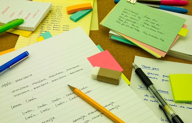 Paper, notepads, pens and erasers are spread out on a table. Various words in both English and Spanish are written on the pads.