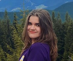 Anna has long hair brown and blue eyes. She is standing outdoors, there is a forest and a range of mountains behind her. She is wearing a purple T-shirt.