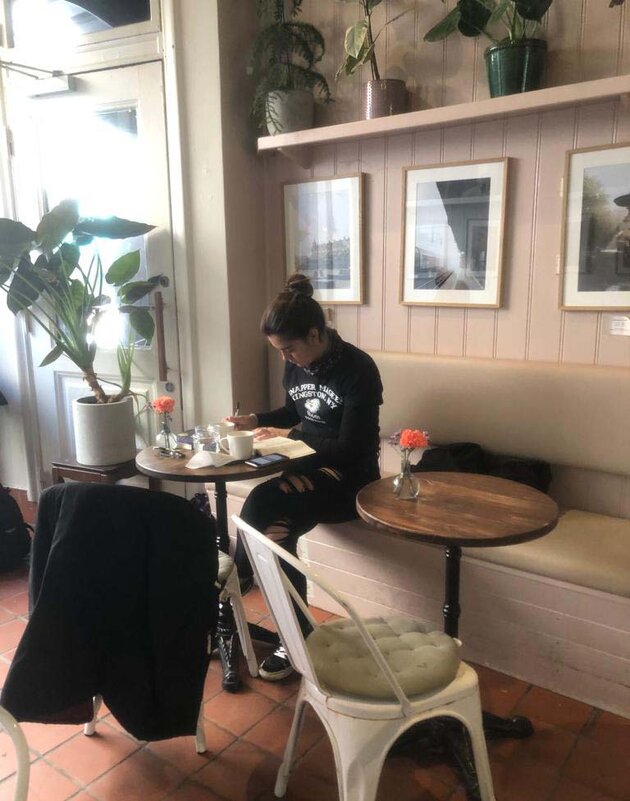 nne på ett café med ljus träpanel och tavlor på väggarna sitter Raquel vid ett bord och skriver i en anteckningsbok. Hon har det mörka håret uppsatt i en knut och är klädd i svarta byxor med hål på benen och en svart tröja med vit text.