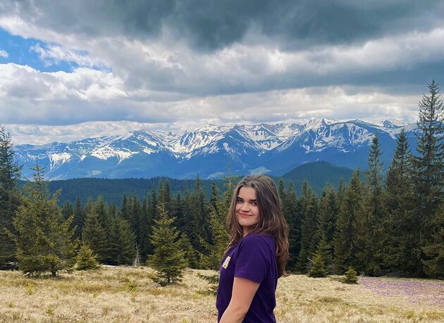 Anna has long hair brown and blue eyes. She is standing outdoors, there is a forest and a range of mountains behind her. She is wearing a purple T-shirt.