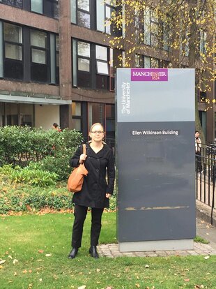 Lina Sturfelt is standing on campus next to a sign saying “Ellen Wilkinson Building”. She is wearing black trousers and a black coat and carrying a brown shoulder bag. She has light-coloured hair styled up in a bun and glasses. The photo was taken on the University of Manchester campus.