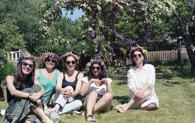 Fem kvinnor med solglasögon och blomsterkransar i håret sitter på en gräsmatta. Bakom dem står en låg majstång med massor av gröna blad och lila blommor i. Det firas midsommar!