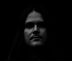 A black and white close-up of Jakob looking at the camera with a serious expression. His long, straight hair moves slightly in the breeze and is pulled to one side. Jakob is wearing a dark shirt.