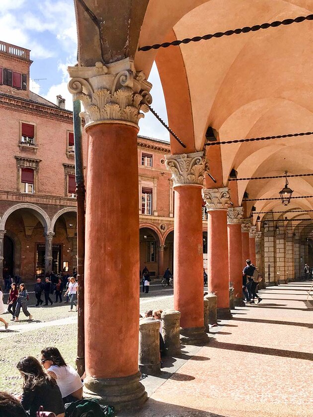 En stadsbild från Bologna. Till höger i bilden syns rödaktiga kolonner som håller upp ett valvtak, och nedanför dem sitter människor på stenavsatser i solen. Utanför på gatan till vänster passerar gående förbi, och på andra sidan gatan syns byggnader med liknande kolonner och gallerior.
